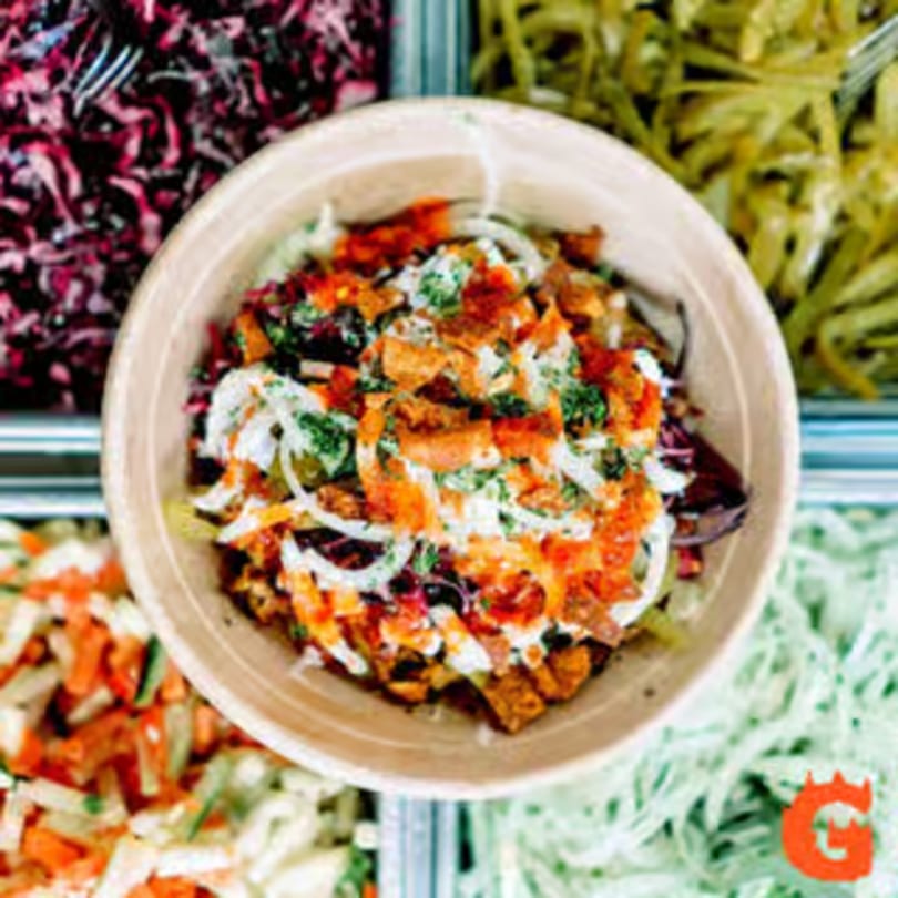 Top-down view of a colorful Mediterranean bowl with various toppings, surrounded by other fresh ingredients.